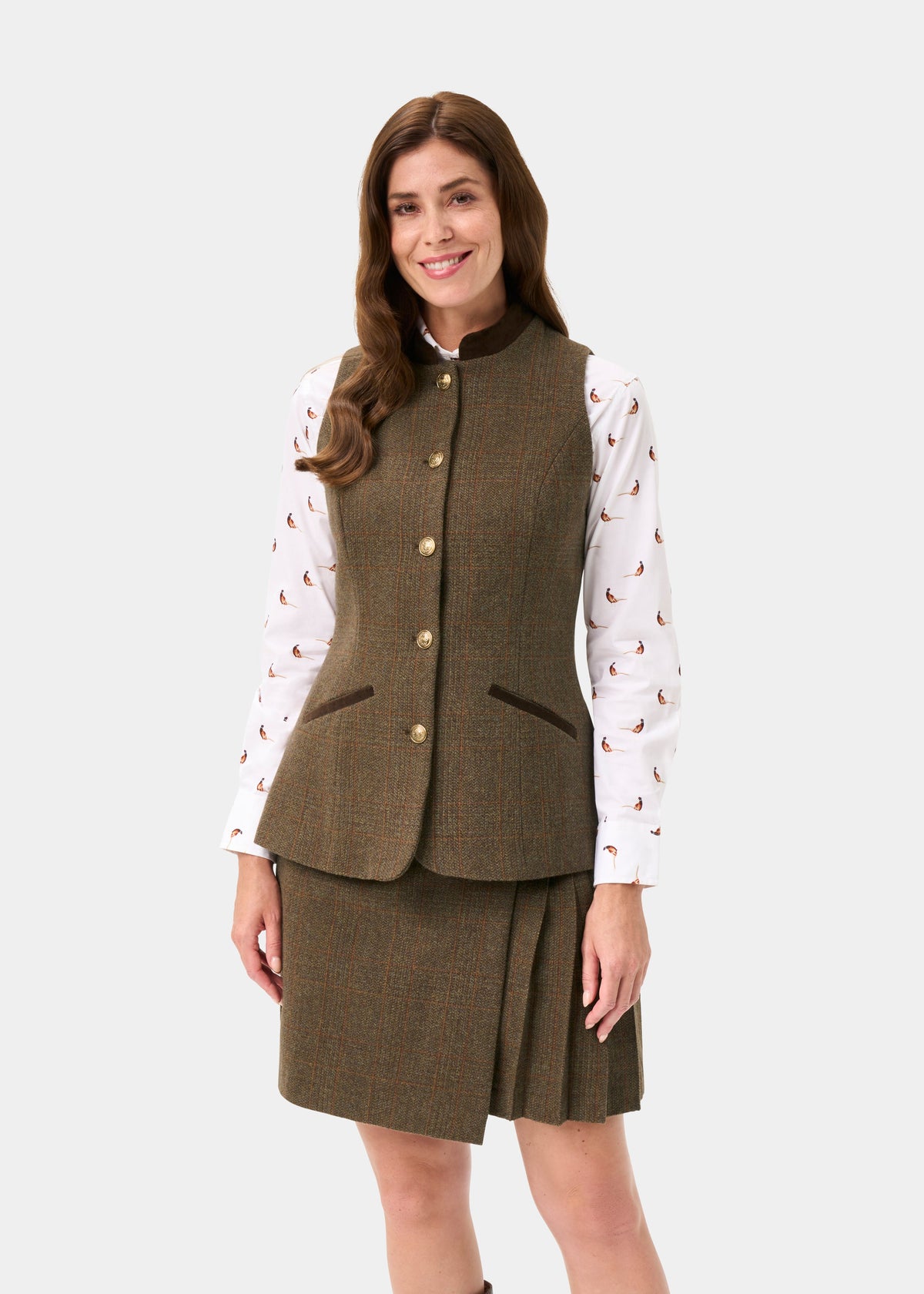 A woman modeling a brown tweed vest and matching pleated skirt set, worn over a white long-sleeve shirt with a small paisley print, against a light gray studio background.