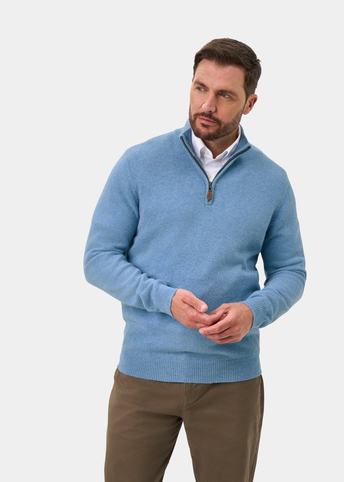 A man wears a Sutherland Lambswool Quarter Zip Jumper In Glacier over a white collared shirt and brown trousers.
